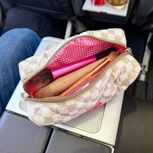 May include: A quilted beige and white checkered makeup bag with a zipper closure. The interior is lined with a pink and white gingham fabric. Inside the bag are two makeup brushes, one with a bright pink handle and one with a tan handle.