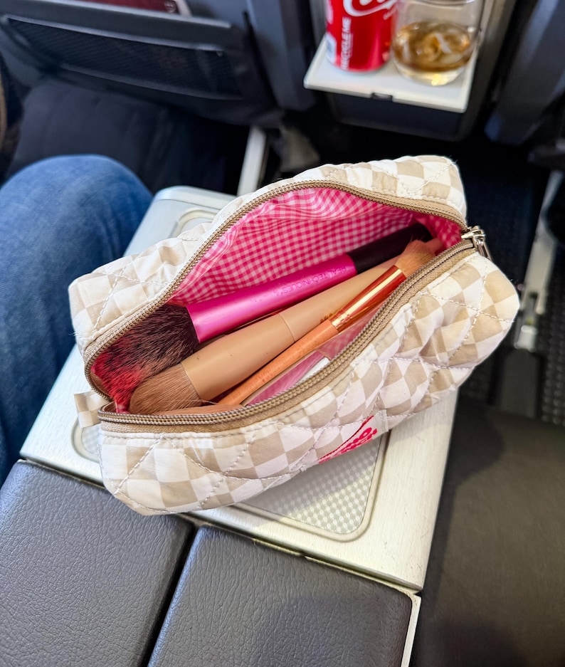 May include: A quilted beige and white checkered makeup bag with a zipper closure. The interior is lined with a pink and white gingham fabric. Inside the bag are two makeup brushes, one with a bright pink handle and one with a tan handle.