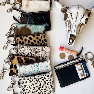 May include: A stack of nine cowhide keychains with different patterns, including leopard print, turquoise, and brown and white spots. Each keychain has a silver key ring attached. A small pink lip balm tube and two silver coins are visible in the background.