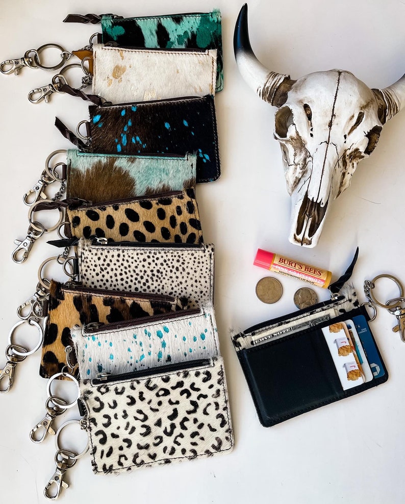 May include: A stack of nine cowhide keychains with different patterns, including leopard print, turquoise, and brown and white spots. Each keychain has a silver key ring attached. A small pink lip balm tube and two silver coins are visible in the background.