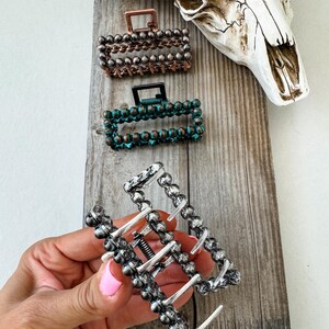 May include: Rectangular hair claw clips in copper, teal, and silver tones. The clips are adorned with small, round beads and feature a spring mechanism. The image also includes a cow skull on a wooden surface.