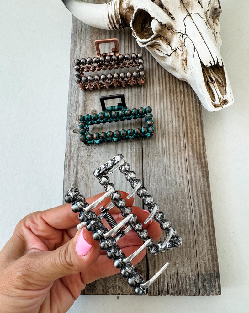 May include: Rectangular hair claw clips in copper, teal, and silver tones. The clips are adorned with small, round beads and feature a spring mechanism. The image also includes a cow skull on a wooden surface.