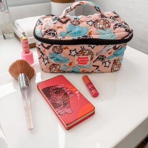 May include: A peach-colored quilted makeup bag with a black zipper and handle, featuring a pattern of disco balls, cowboy hats, and stars. A makeup brush, lipstick, and a small book are also visible.