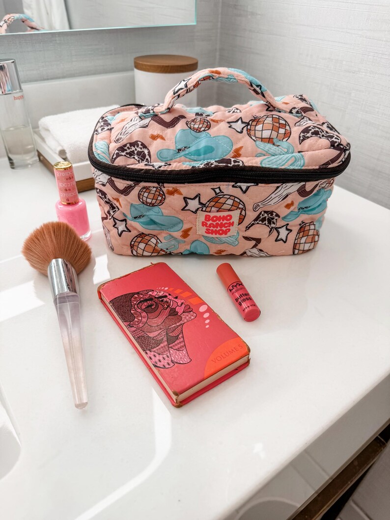 May include: A peach-colored quilted makeup bag with a black zipper and handle, featuring a pattern of disco balls, cowboy hats, and stars. A makeup brush, lipstick, and a small book are also visible.