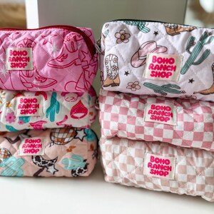 May include: Six quilted zippered pouches stacked in two columns. The pouches feature various patterns including cowboy boots, cacti, flowers, stars, and checkered prints in shades of pink, white, and tan. Each pouch has a label reading BOHO RANCH SHOP.