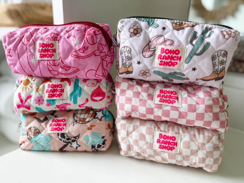 May include: Six quilted zippered pouches stacked in two columns. The pouches feature various patterns including cowboy boots, cacti, flowers, stars, and checkered prints in shades of pink, white, and tan. Each pouch has a label reading BOHO RANCH SHOP.
