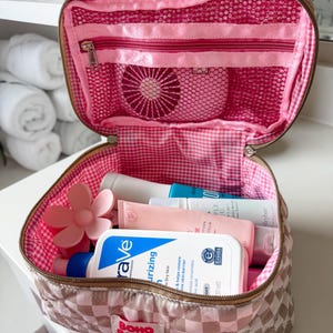 May include: Open pink and brown quilted cosmetic bag with a gingham interior. The bag contains various skincare products, a pink flower-shaped item, and a mesh pocket. The bag has a zipper closure and a "BOHO RANCH SHOP" label.