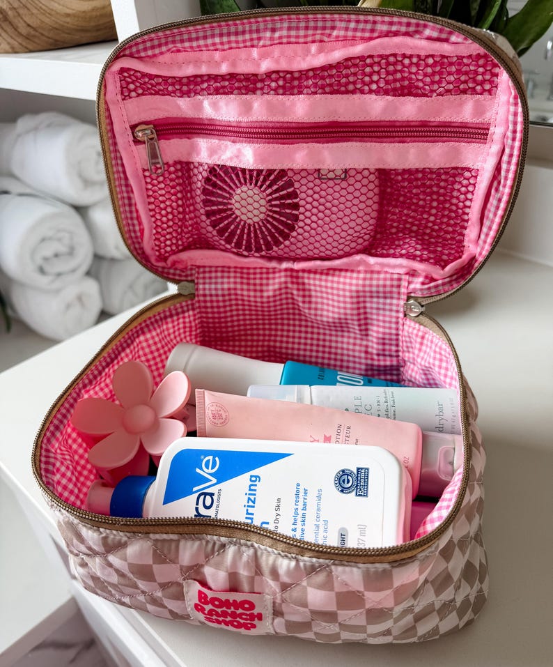 May include: Open pink and brown quilted cosmetic bag with a gingham interior. The bag contains various skincare products, a pink flower-shaped item, and a mesh pocket. The bag has a zipper closure and a "BOHO RANCH SHOP" label.