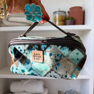 May include: A rectangular cosmetic bag made of patchwork cowhide in shades of turquoise, brown, and white. The bag features a dark brown zipper, a top handle, and a tan leather patch with the text Boho Ranch Shop and a cactus graphic.