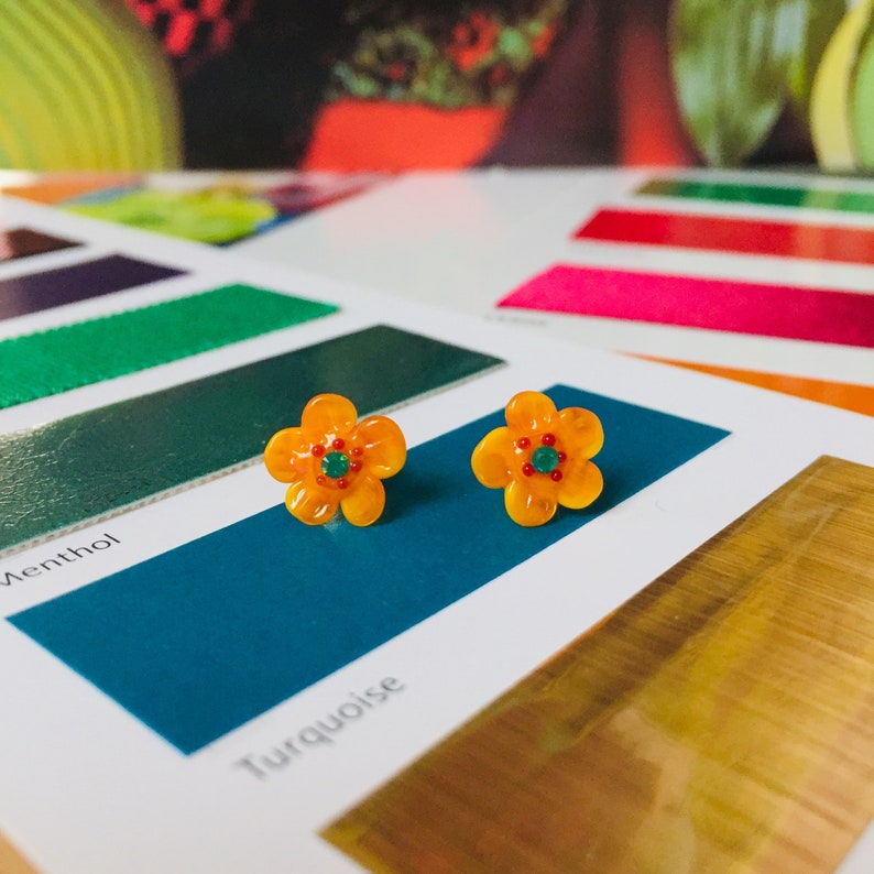 Puede incluir: Un par de pendientes de bot&oacute;n con forma de flor. Cada pendiente presenta un dise&ntilde;o naranja brillante de cinco p&eacute;talos con un peque&ntilde;o centro verde redondo. Los pendientes se colocan sobre un fondo de muestras de color, incluyendo turquesa y oro.