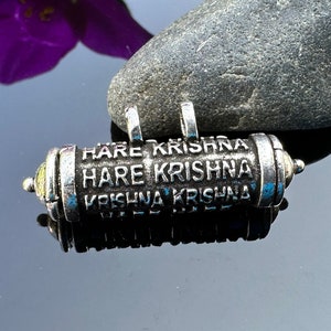 Pode incluir: Um amuleto de prata com as palavras "Hare Krishna" repetidas em cada lado. O amuleto &eacute; cil&iacute;ndrico e tem um acabamento liso e polido.