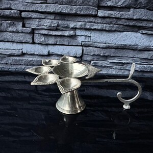 May include: A silver metal diya with five wicks and a handle. The diya is designed in a traditional Indian style.