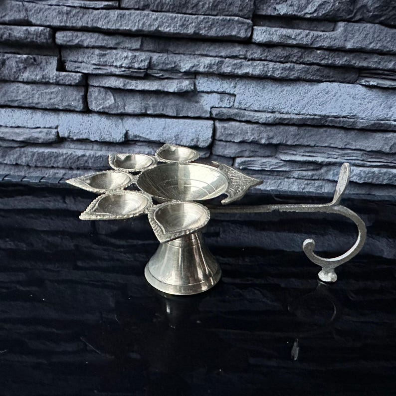 May include: A silver metal diya with five wicks and a handle. The diya is designed in a traditional Indian style.