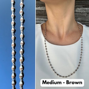 Puede incluir: Un collar de cadena de plata con cuentas de madera marr&oacute;n. El collar est&aacute; puesto en un maniqu&iacute; que lleva una camisa blanca. El texto "Medium - Brown" est&aacute; en la parte inferior de la imagen.