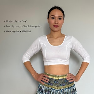 May include: A white, three-quarter sleeve crop top with a scoop neckline. The top is worn with a patterned skirt. The image includes text that reads: Model: 5'5", Bust: 32.7" at fullest point, and Wearing size XS (White).
