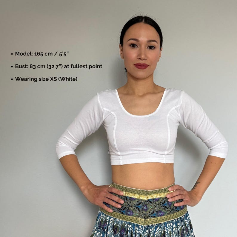 May include: A white, three-quarter sleeve crop top with a scoop neckline. The top is worn with a patterned skirt. The image includes text that reads: Model: 5'5", Bust: 32.7" at fullest point, and Wearing size XS (White).