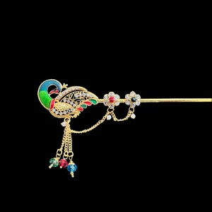 Puede incluir: Un accesorio para el cabello de color dorado con un diseño de pavo real con detalles de esmalte azul, verde y rojo. El accesorio tiene un alfiler largo y recto y cadenas colgantes con cuentas de colores. El pavo real está adornado con cristales transparentes.