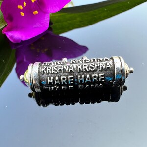 Pode incluir: Um amuleto de metal prateado com as palavras "Hare Krishna Krishna Hare Hare" gravadas nele.