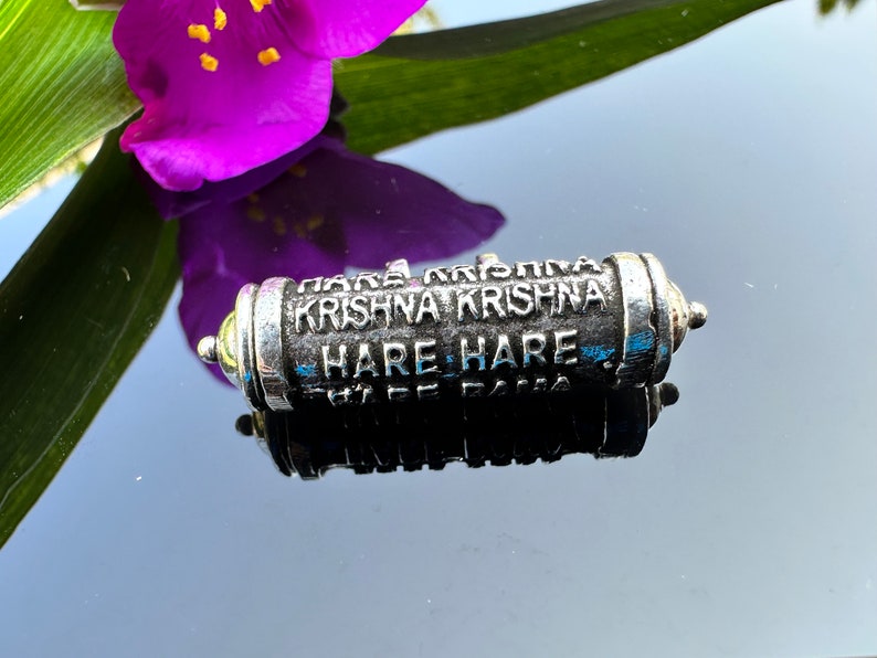 Pode incluir: Um amuleto de metal prateado com as palavras "Hare Krishna Krishna Hare Hare" gravadas nele.