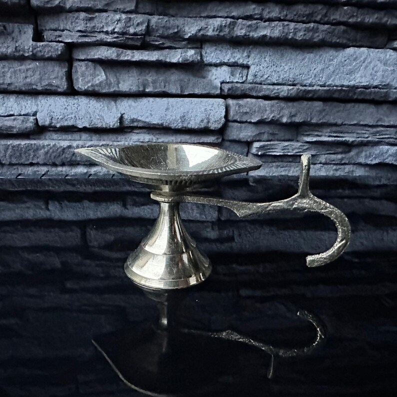 May include: A silver metal diya with a handle, used for burning oil or ghee as a religious offering.