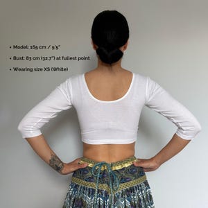 May include: A white, three-quarter sleeve crop top with a scoop neckline. The model is wearing size XS. The bust measurement is 32.7 inches at the fullest point. The model is 5'5" tall. The text on the image says: Model: 5'5", Bust: 32.7", Wearing size XS (White).