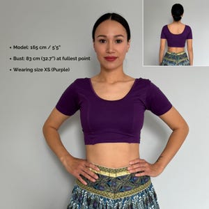 May include: A cropped, short-sleeved purple top with a scoop neckline. The model is wearing size XS. The image also includes a smaller image of the back of the top. The model's bust is 32.7 inches at the fullest point.