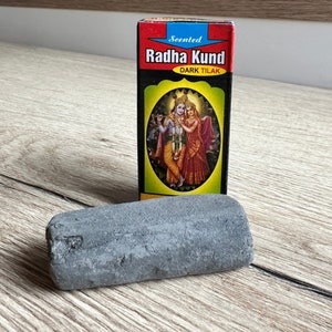 May include: A small, dark gray stone, possibly a grinding stone, sits next to a box of Radha Kund Dark Tilak. The box is red with a yellow border and features a picture of Radha and Krishna.