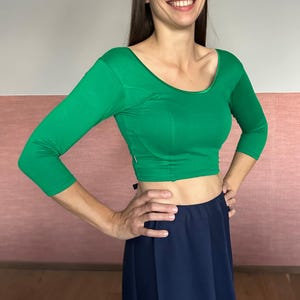 May include: A green, long-sleeved crop top with a scoop neckline. The top is paired with a dark blue skirt. The model is smiling and has their hands on their hips.