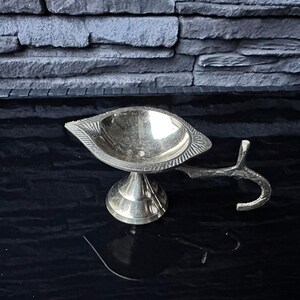 May include: A silver metal diya with a decorative handle. The diya has a round bowl and a raised base.