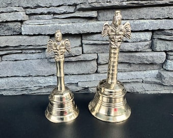 May include: Two gold-colored bells with ornate handles in the shape of a mythical creature. The bells are sitting on a gray stone surface.