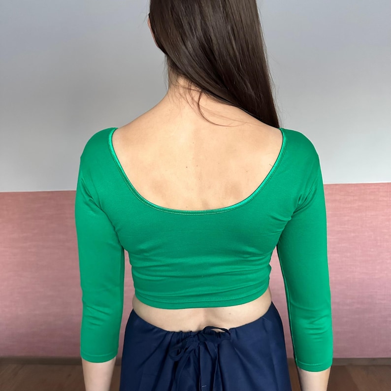 May include: A green, three-quarter sleeve crop top with a scoop neckline. The top is a vibrant green color and appears to be made of a smooth, possibly stretchy fabric. The neckline is trimmed with a darker green material. The model is wearing a navy blue skirt.