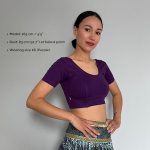 May include: A short-sleeved, purple crop top with a scoop neckline. The top is form-fitting and has a seam detail down the front. The model is wearing size XS. The model's bust is 32.7 inches at the fullest point.