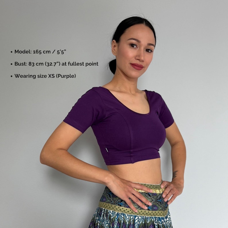 May include: A short-sleeved, purple crop top with a scoop neckline. The top is form-fitting and has a seam detail down the front. The model is wearing size XS. The model's bust is 32.7 inches at the fullest point.
