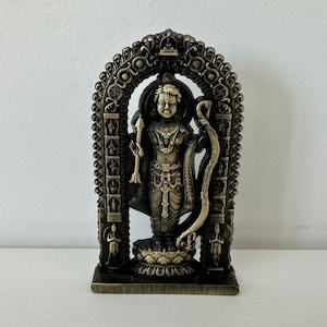 May include: A gold-toned metal statue of a Hindu deity standing in a decorative archway. The statue is detailed with intricate carvings and features a bow and arrow.