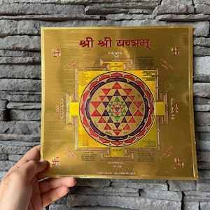 Puede incluir: Placa metálica cuadrada dorada con intrincados diseños geométricos y texto en escritura extranjera. El diseño central presenta un mandala complejo con elementos rojos, morados y verdes. La placa está sostenida por una mano sobre un fondo de piedra texturizada.
