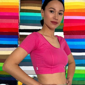 May include: A short-sleeved, hot pink cropped top featuring a scoop neckline with a matching fabric trim. The garment has a fitted silhouette with vertical seam detailing along the front and a small white rectangular label on the side seam.