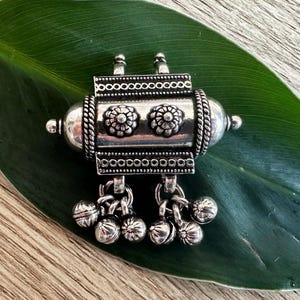 May include: Silver pendant with a cylindrical design, featuring intricate floral patterns and a dangling chain with multiple small bells.