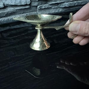 May include: A small, gold-colored brass diya with a handle. The diya has a round base and a shallow bowl for holding oil or ghee.