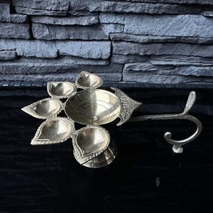 May include: A brass diya with five wicks, each shaped like a teardrop, and a central bowl. The diya has a decorative handle that can be used to hang it.