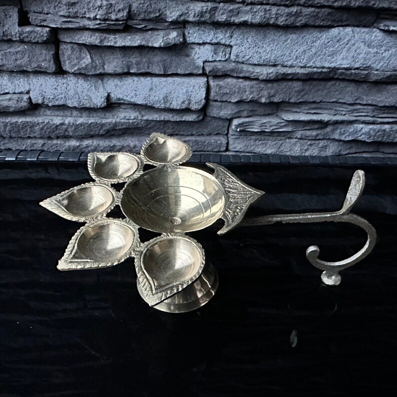 May include: A brass diya with five wicks, each shaped like a teardrop, and a central bowl. The diya has a decorative handle that can be used to hang it.