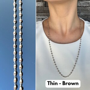 Puede incluir: Un collar de cadena de plata con cuentas marrones. El collar es delgado y tiene un aspecto delicado. El texto "Thin - Brown" es visible en la parte inferior de la imagen.