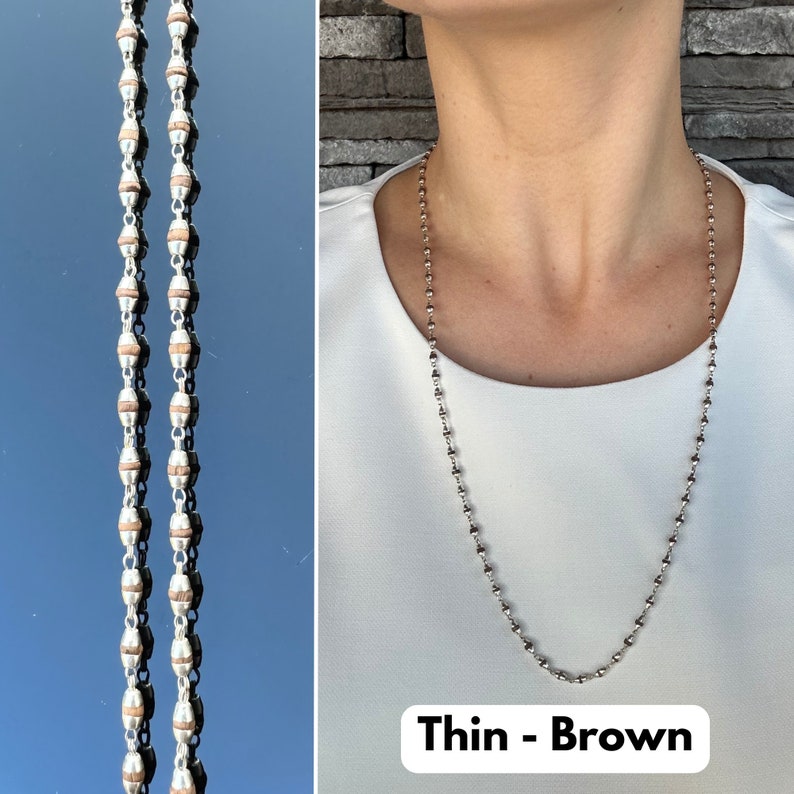 Puede incluir: Un collar de cadena de plata con cuentas marrones. El collar es delgado y tiene un aspecto delicado. El texto "Thin - Brown" es visible en la parte inferior de la imagen.