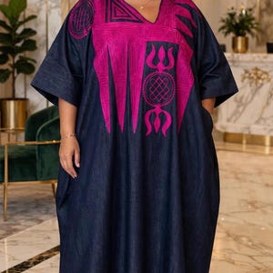 Puede incluir: Un vestido caftán de mezclilla azul oscuro con escote en V y bordado magenta vibrante en el pecho y los hombros. La prenda tiene mangas cortas y anchas, una silueta holgada y se combina con zapatillas deportivas blancas.