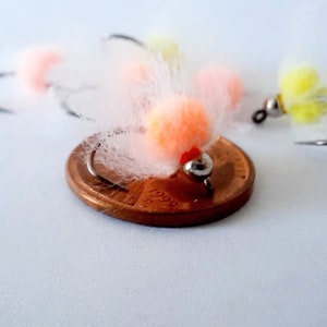 May include: Close-up of a small, white, fluffy fly fishing lure with an orange bead. The lure is sitting on top of a US penny.