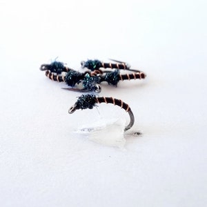 May include: Close-up of several fishing flies. Each fly features a curved hook with a dark, wrapped body and a tuft of dark green material. The flies are arranged on a white surface, with one fly in sharp focus.