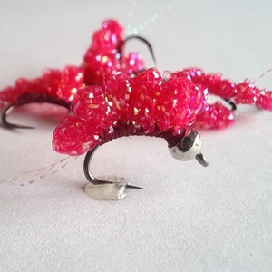 May include: Close-up of three pink fly fishing lures with iridescent sparkle. The lures are tied on hooks with a silver bead on the front.