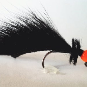 May include: Black fly fishing lure with an orange bead. The lure has a long, black, fuzzy body.