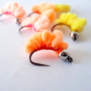 May include: A close-up of six colorful fly fishing lures. The lures are made of fluffy material and have a bead on the hook. The colors of the lures are orange, yellow, and pink.