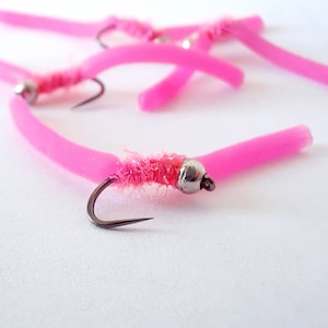 May include: Five pink rubber worm fishing lures with a fuzzy pink body and a silver bead. The lures are tied to a black hook.