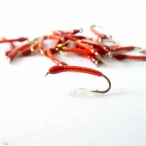 May include: A close-up of a pile of red fishing lures with gold hooks. The lures are made of a soft, flexible material and are designed to resemble worms.